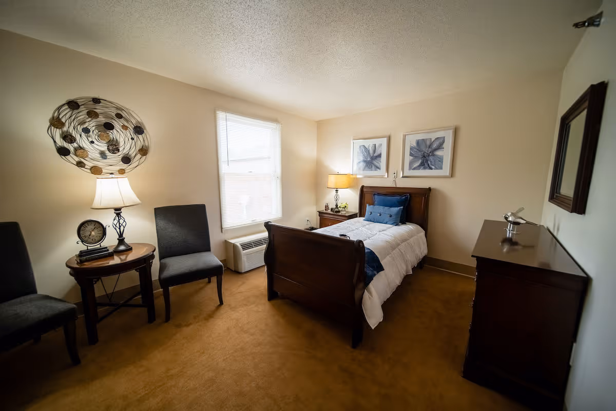 A cozy bedroom with a single wooden bed dressed in white bedding and blue pillows. The room features two gray chairs and a round wooden side table with a lamp and clock. There is a window with blinds, two framed floral artworks above the bed, a wooden dresser with a decorative item, and a wall mirror. The carpet is brown and the walls are light-colored.