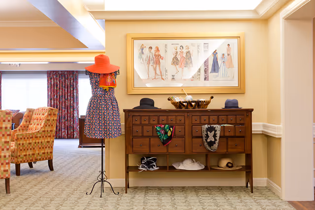 Decorated common area with a mannequin wearing a dress and hat, a wooden console table displaying hats and accessories, and a framed fashion print above.
