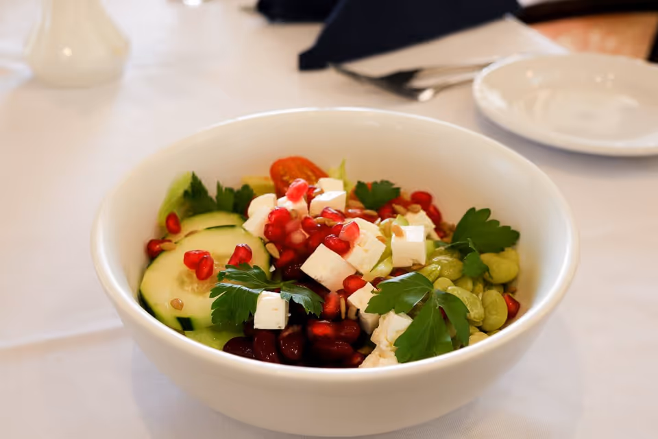 A white bowl filled with a fresh salad containing cucumber slices, pomegranate seeds, kidney beans, feta cheese cubes, green beans, and parsley leaves, placed on a white tablecloth with a plate and napkin in the background.