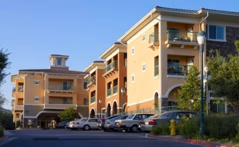 Exterior view of a multi-story senior living building with balconies and parked cars.