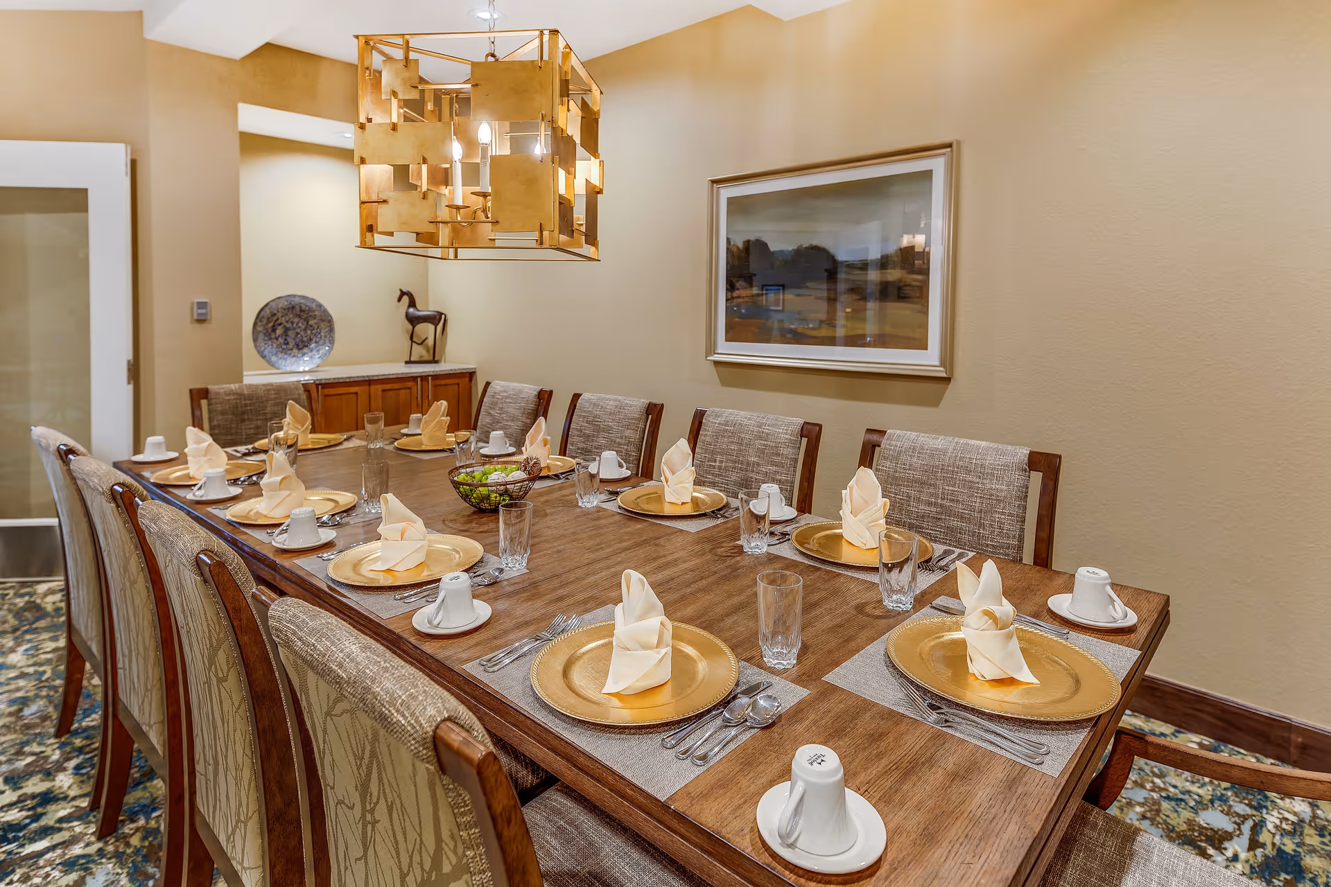 A formal dining room with a long wooden table set for ten people. Each place setting includes a gold charger plate, a folded beige napkin, a glass, silverware, and a white cup with a saucer. The room has beige walls, a modern gold chandelier hanging above the table, a framed landscape painting on the wall, and a sideboard with decorative items including a plate and a horse figurine.