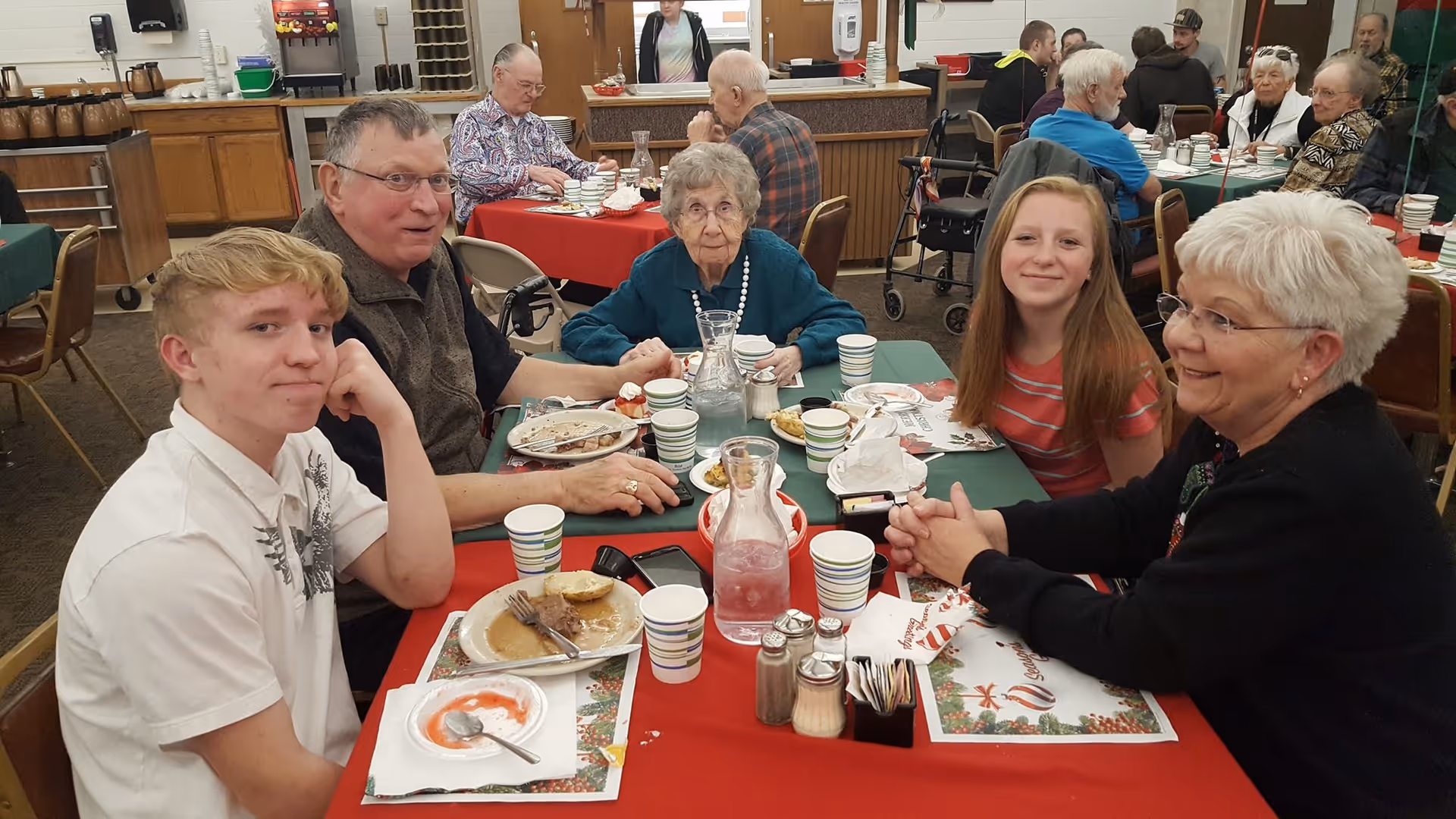 A group of five people, including elderly individuals and younger adults, sitting around a table with plates of food and cups in a communal dining area. The table is covered with a red and green tablecloth with holiday-themed placemats. Other people are visible dining in the background.