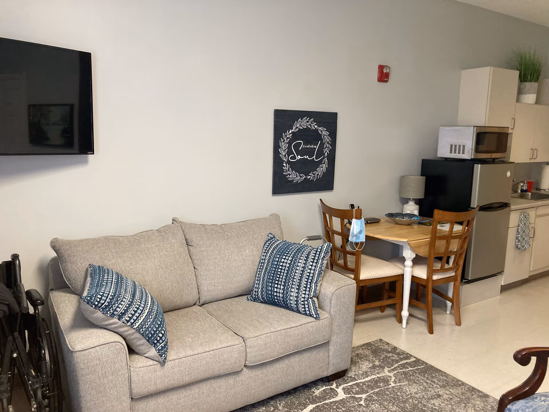 A small living area with a gray couch adorned with two blue patterned pillows. To the right, there is a wooden table with two chairs, a lamp, and a face mask hanging on one chair. Behind the table is a compact kitchen area with a microwave, mini refrigerator, sink, and cabinets. A flat-screen TV is mounted on the wall to the left, and a decorative wall art piece with a wreath and the word 'Soul' is hung above the table.