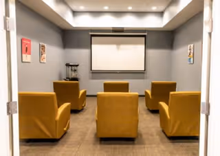 A small theater room with five mustard yellow armchairs arranged in two rows facing a blank projector screen on a gray wall. The room has gray walls, a brown carpeted floor, recessed ceiling lights, and framed pictures on the side walls. A popcorn machine is placed on a small table to the left of the screen.