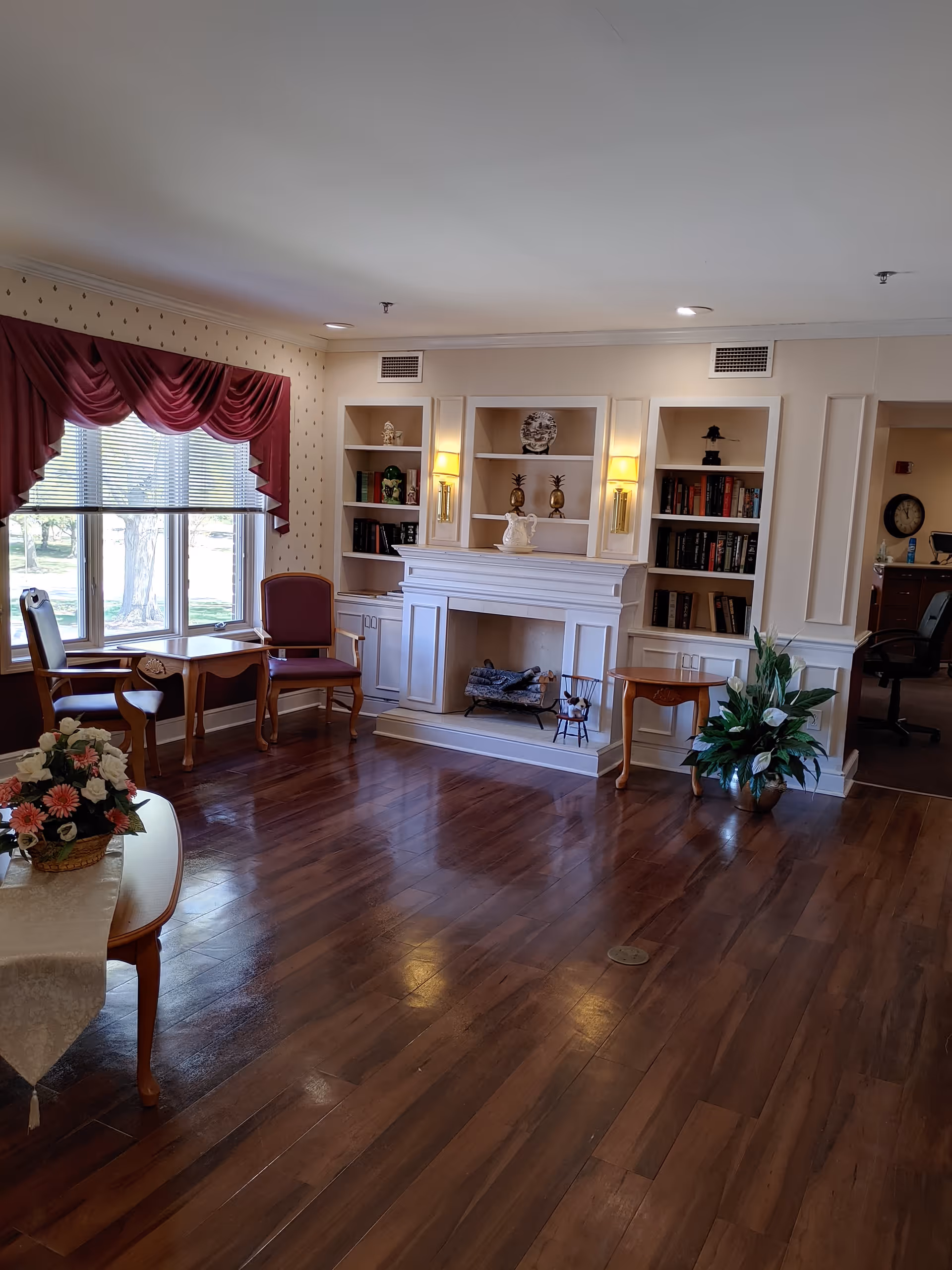A cozy living room area with polished wooden floors, a white fireplace with built-in shelves on either side holding books and decorative items, two wall sconces with lights, a large window with red drapes, wooden chairs and tables, a floral arrangement on a table with a white tablecloth, and a potted plant near the fireplace.