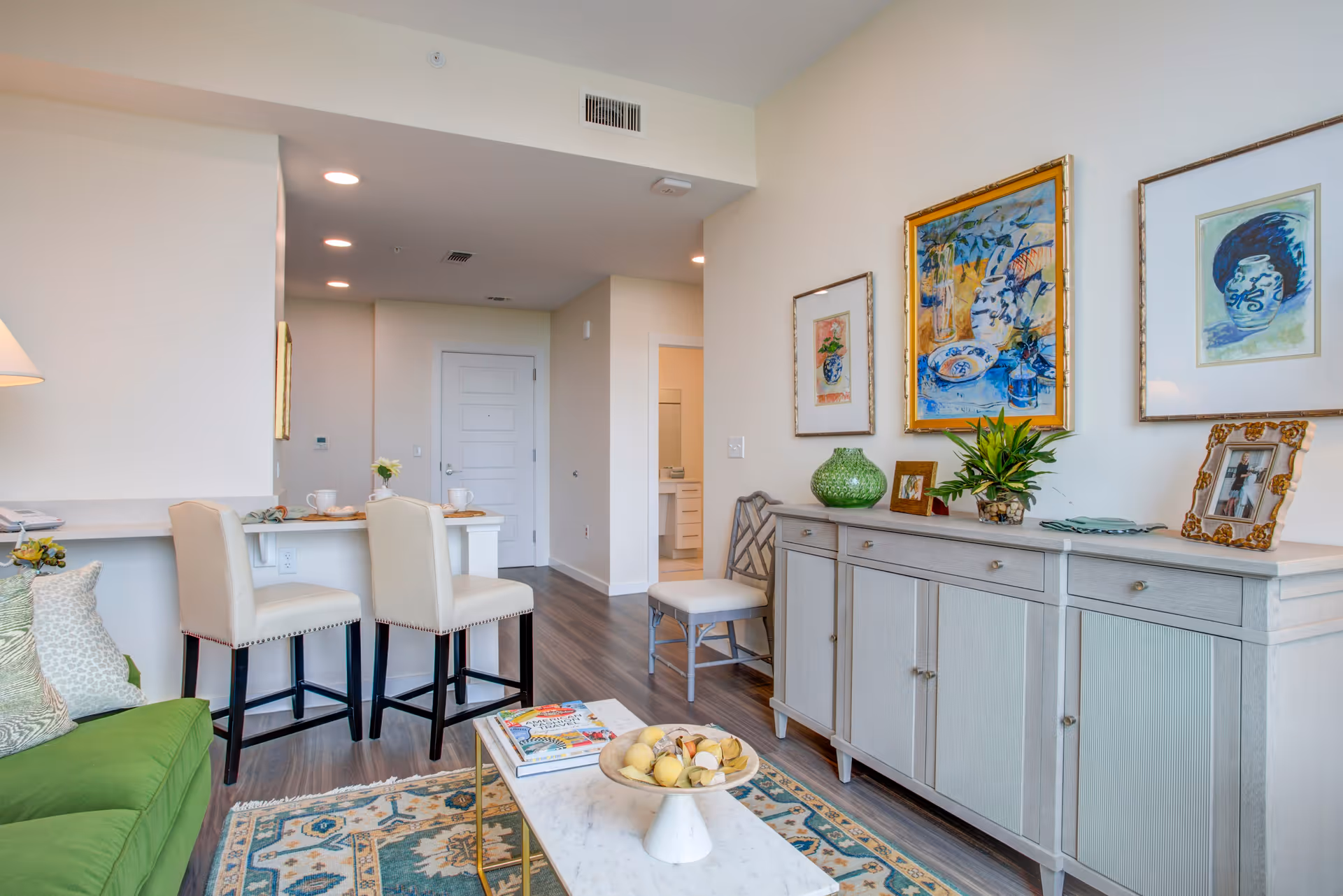 A bright and modern living room area with a green sofa on the left, two beige bar stools at a kitchen counter, a white sideboard with decorative items and framed artwork on the wall above it, a small chair next to the sideboard, and a marble coffee table with a bowl of fruit and magazines on a patterned rug.
