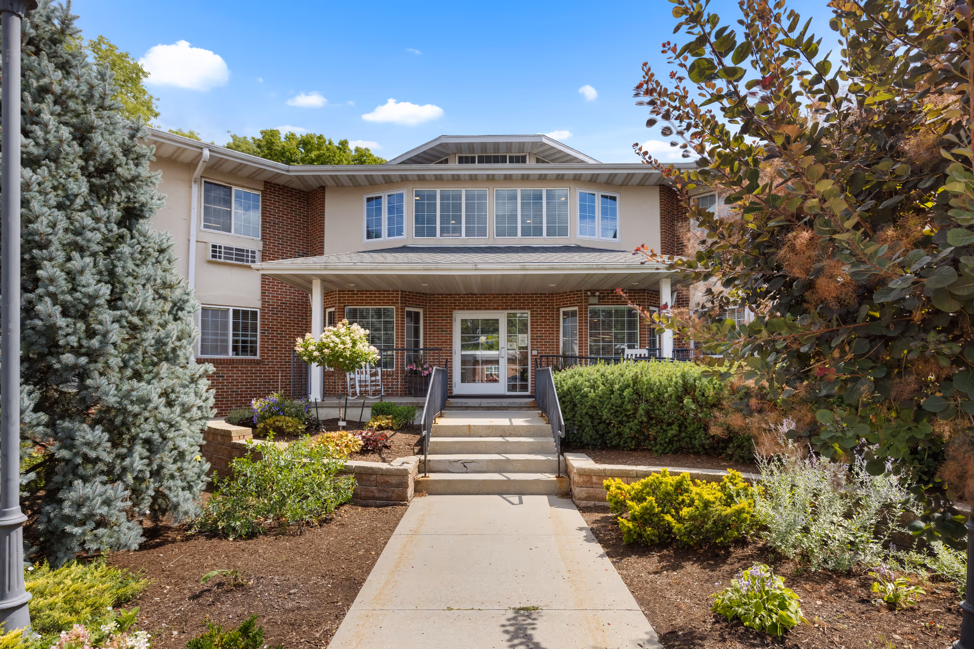 Front exterior view of a two-story assisted living facility building with a covered entrance, steps leading up to the door, and landscaped garden beds with various shrubs and trees on either side of the walkway.