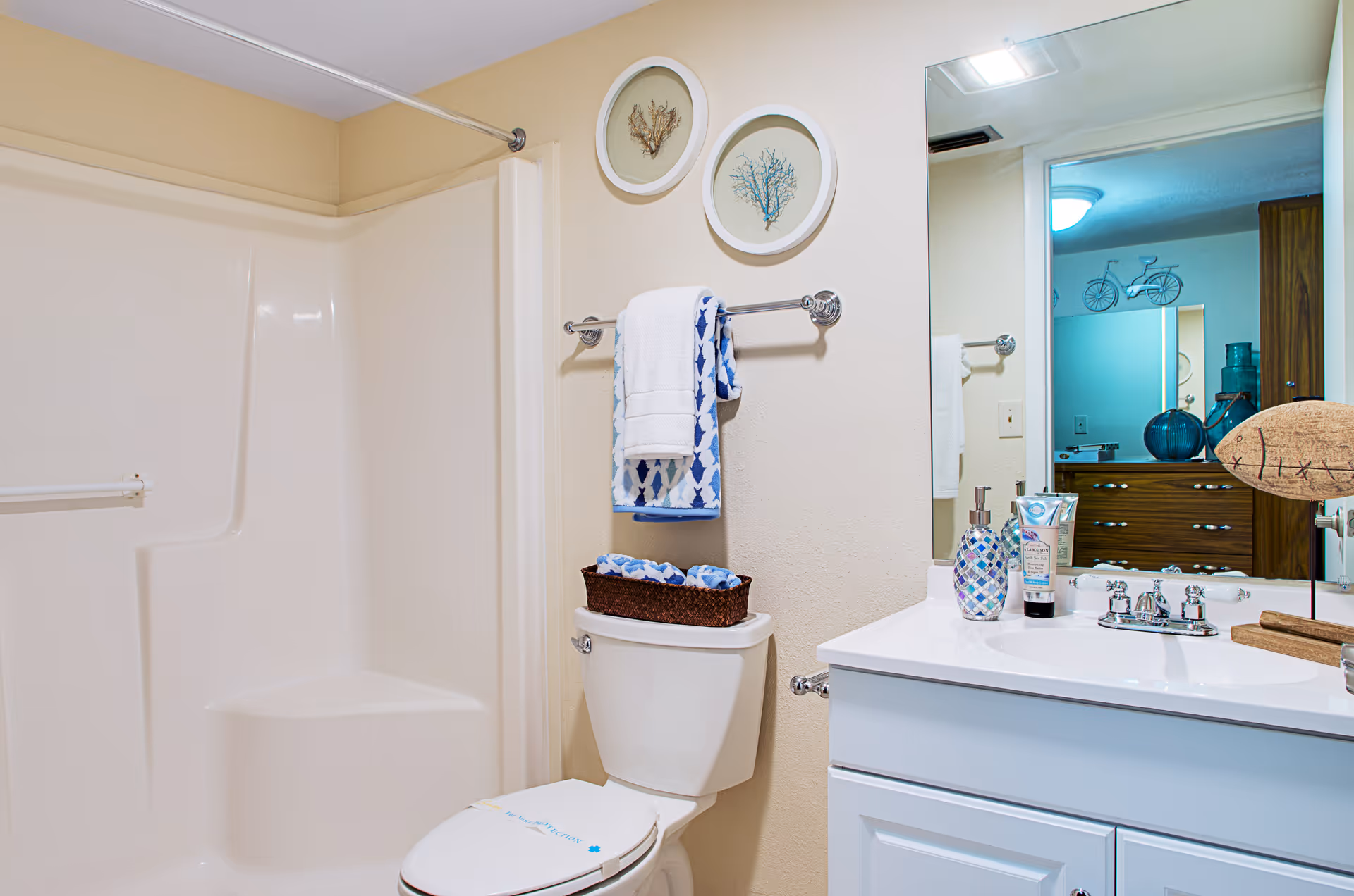 A clean bathroom featuring a white shower with a built-in seat, a toilet with a basket of towels on top, a towel rack with folded towels, a white vanity with a sink, soap dispenser, and lotion, and a large mirror reflecting part of an adjacent room with wooden furniture and decorative items.