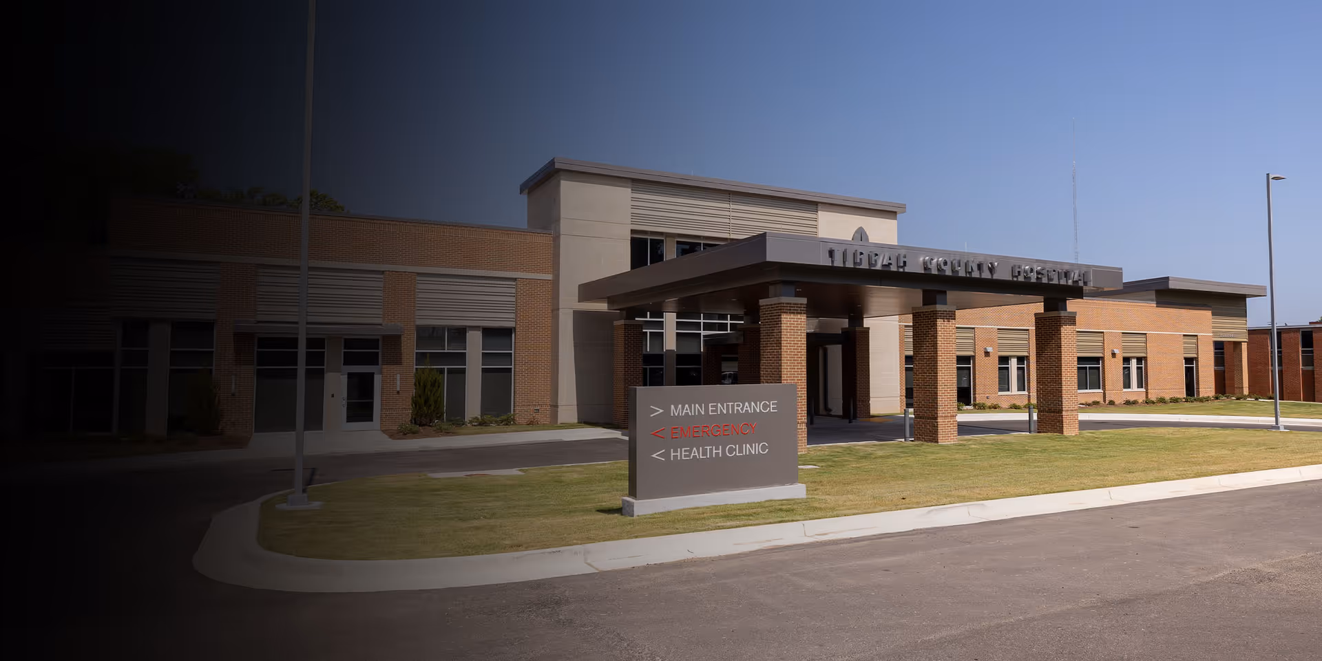 Exterior view of Tippah County Hospital building with a covered entrance supported by brick columns. There is a sign in front indicating directions to the main entrance, emergency, and health clinic. The sky is clear and the surrounding area is landscaped with grass.