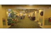 Carpeted communal dining and lounge area with tables, chairs, plants, and residents in a senior living facility.