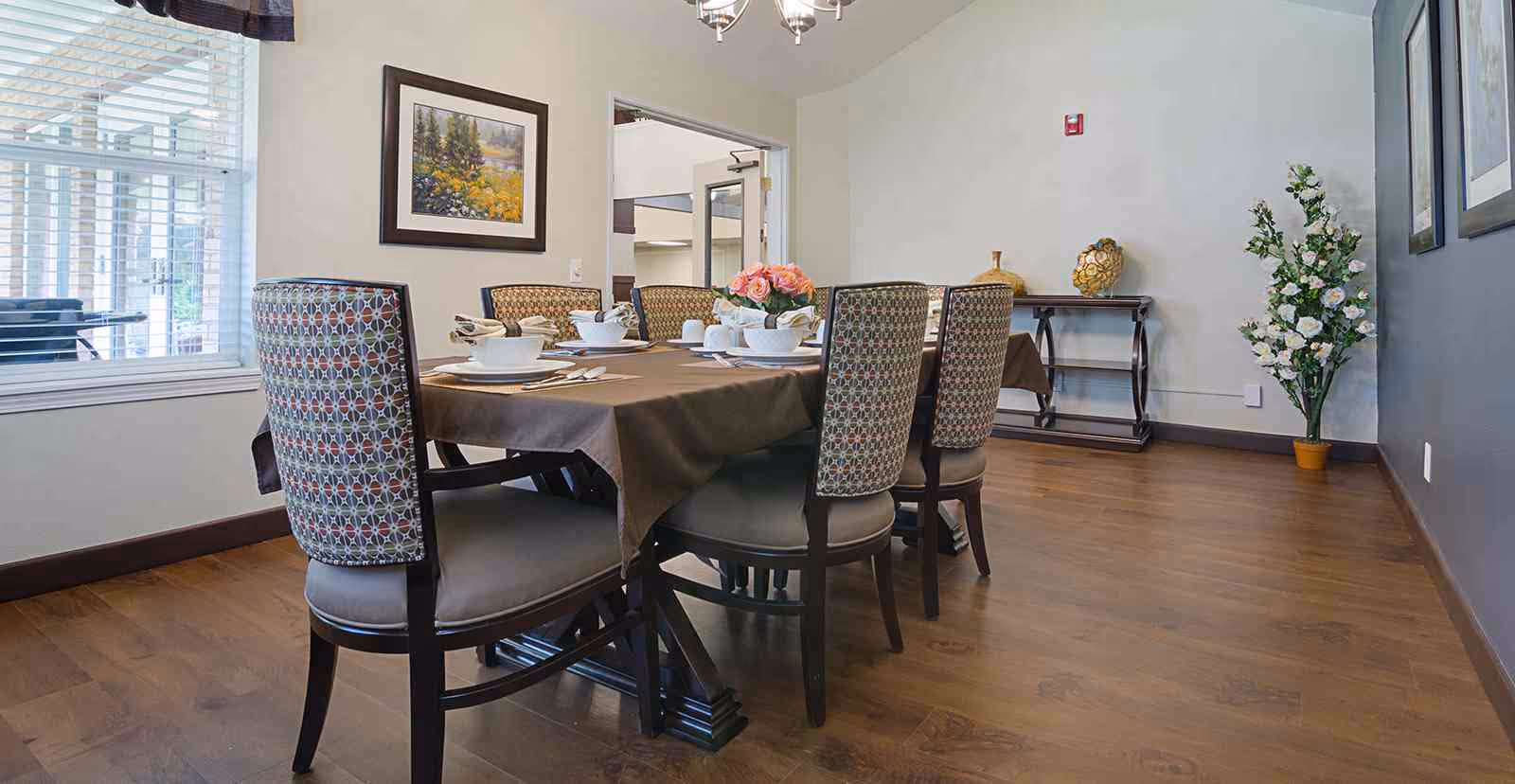 Dining room with a rectangular table set for six, patterned upholstered chairs, hardwood flooring, and decorative wall art and plants.