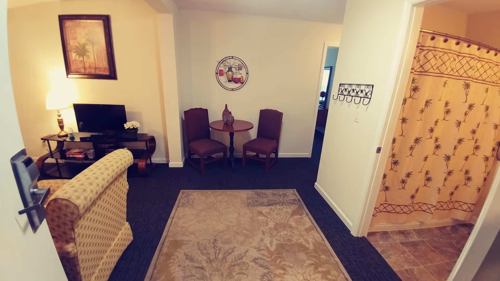 Interior view of a senior living apartment showing a small living area with a patterned armchair, a TV on a stand with a lamp and framed artwork above it, a round table with two chairs against the wall, and a bathroom with a shower curtain featuring a palm tree design.