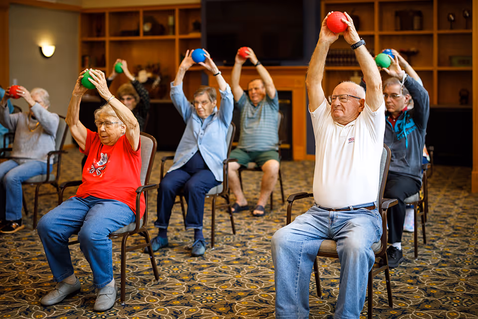 A group of elderly people seated in chairs in a spacious room, participating in a seated exercise class. They are holding colorful balls above their heads, stretching their arms. The room has patterned carpet, wooden shelves, and a television mounted on the wall.