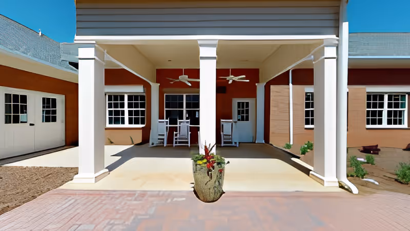 Covered outdoor patio area with white columns, ceiling fans, and white rocking chairs. The patio is attached to a building with brown walls and white-framed windows. There is a planter with flowers in the center foreground and a clear blue sky above.