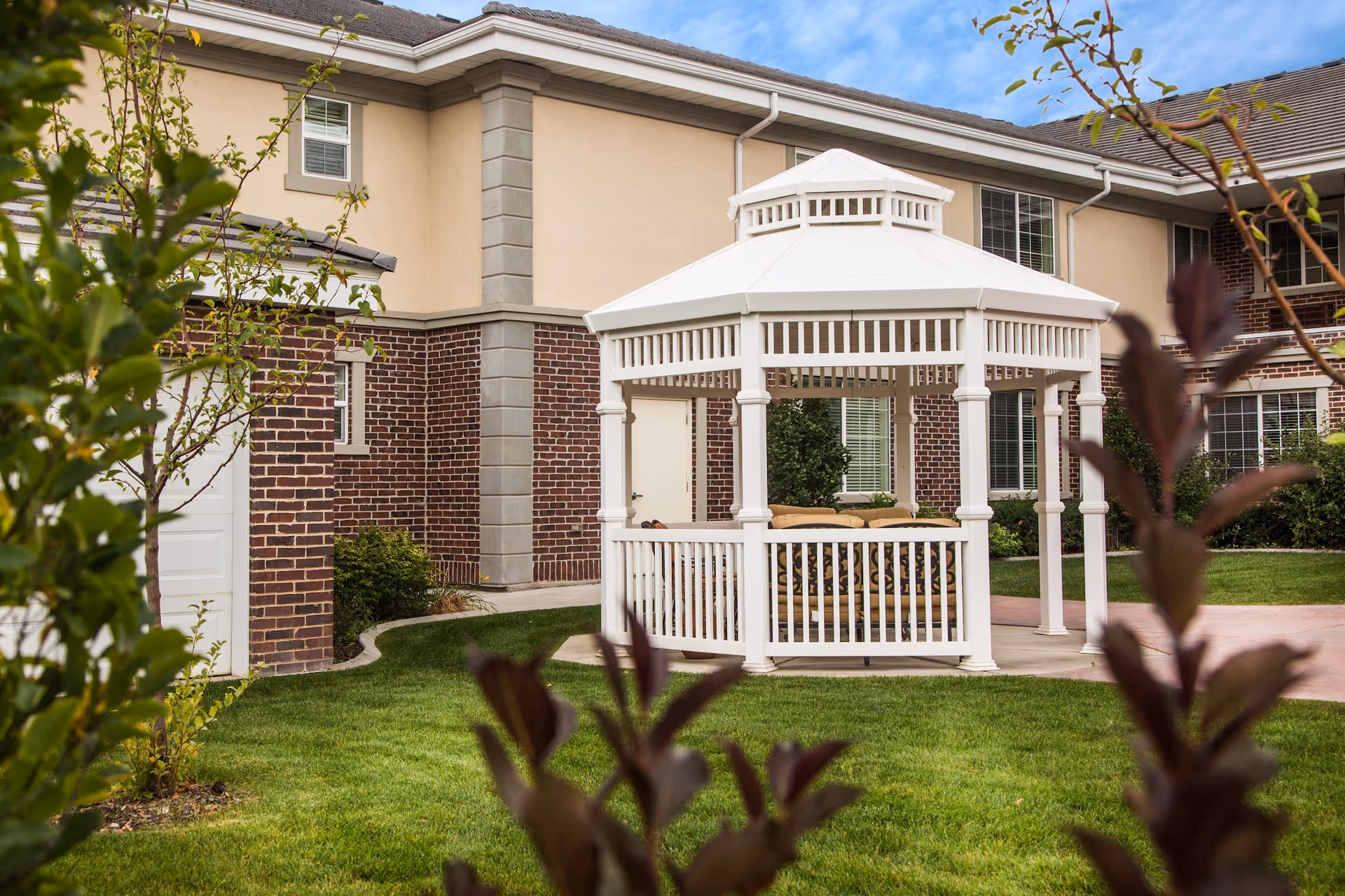 A white gazebo with cushioned seating inside is situated on a well-maintained lawn in front of a two-story building with brick and beige exterior walls and multiple windows under a partly cloudy sky.