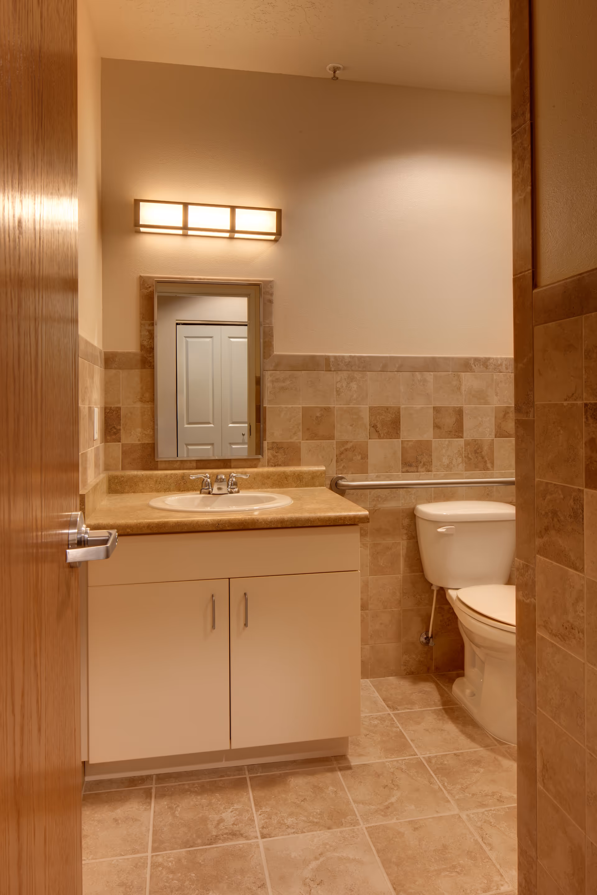 A clean bathroom with beige tiled walls and floor, featuring a white sink with a tan countertop and cabinet below. Above the sink is a rectangular mirror with a modern light fixture. To the right is a white toilet with a metal grab bar on the wall beside it.