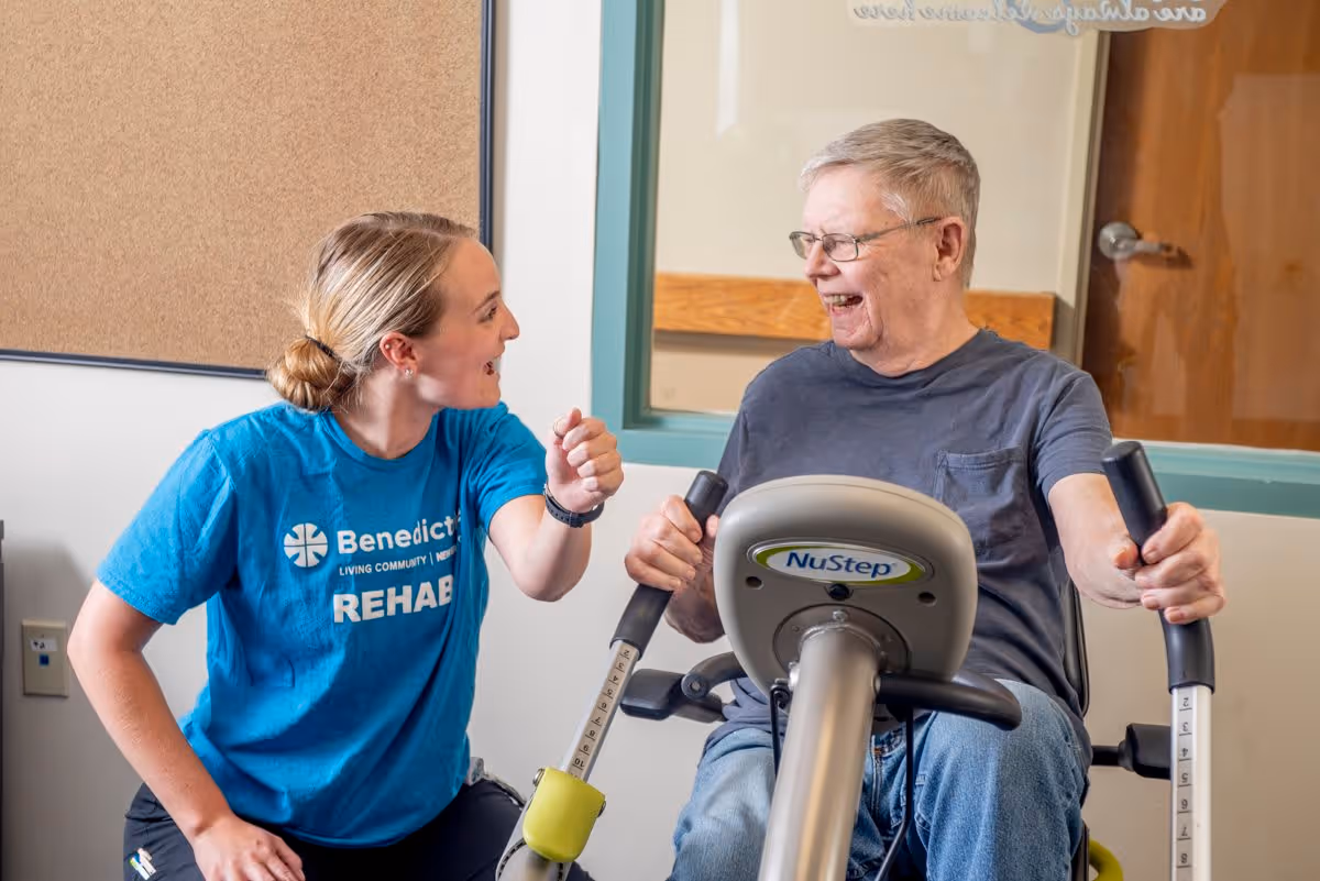 A female rehab staff member wearing a blue Benedictine Living Community-Anoka shirt encourages and smiles at an elderly man who is exercising on a NuStep machine in a rehab or therapy room.