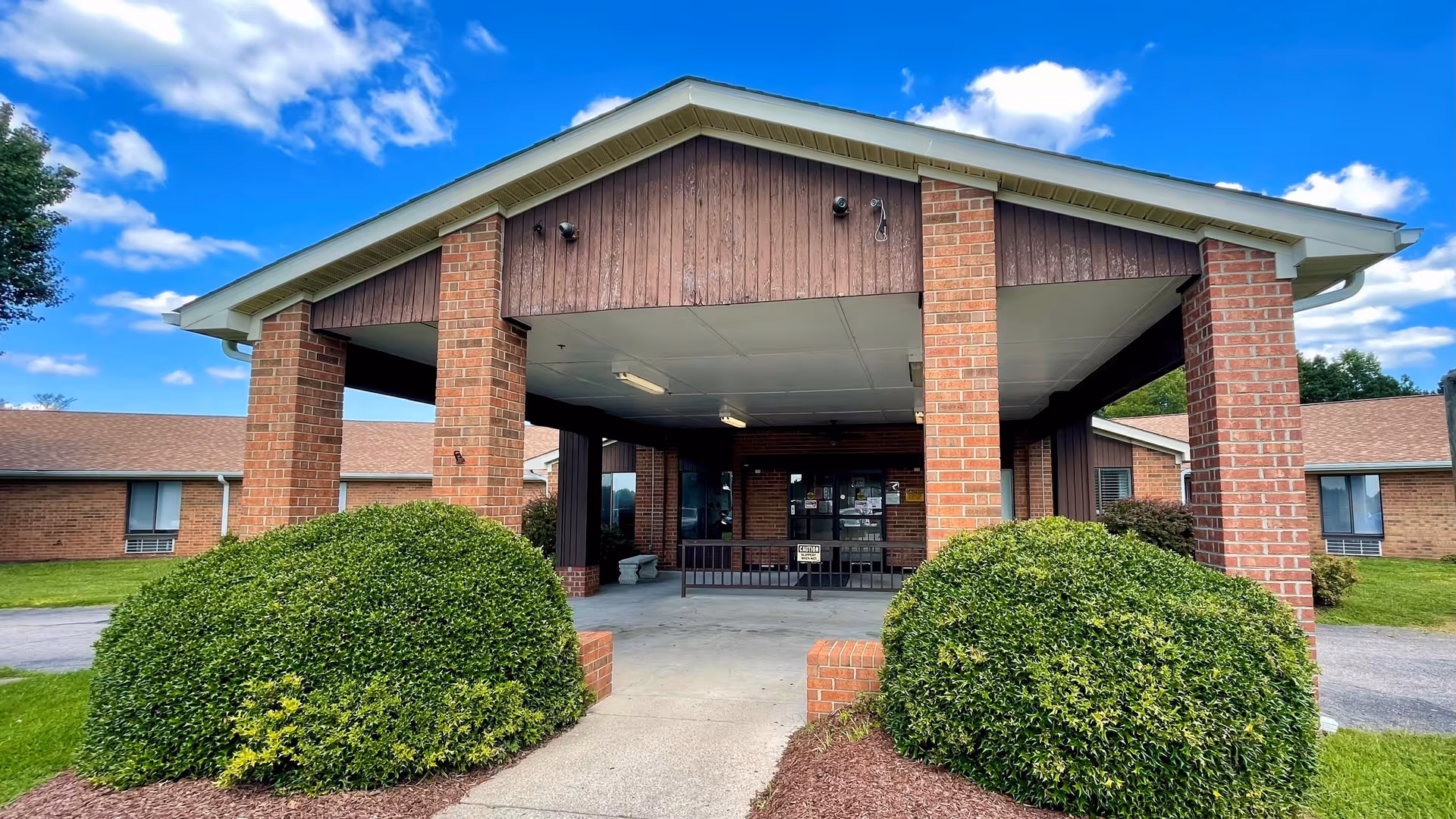 Front entrance of a brick building with a covered driveway supported by four brick pillars, surrounded by green bushes and grass under a blue sky with scattered clouds.