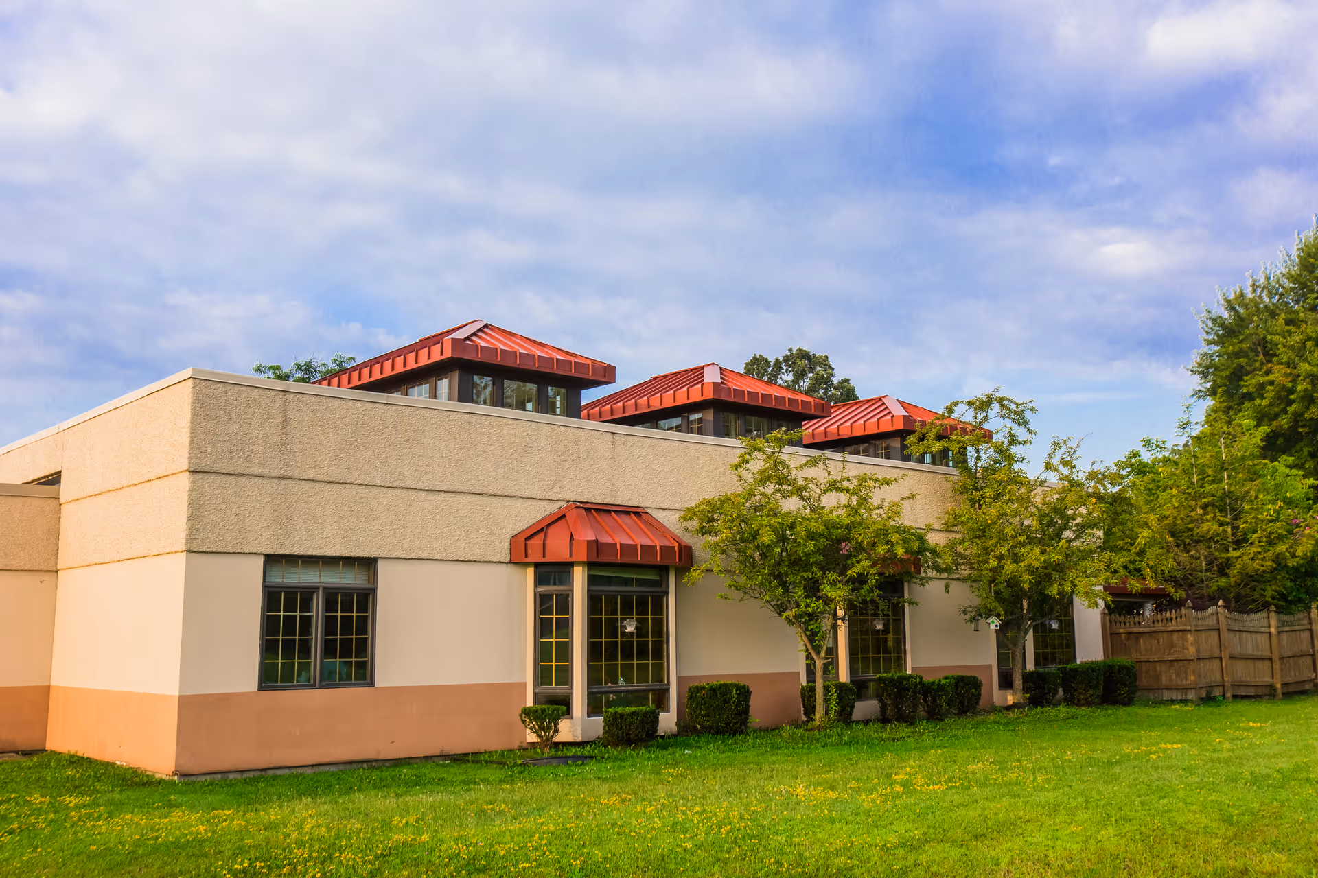 Exterior view of Northgate Health Care Facility showing a beige and peach-colored building with red metal awnings over the windows, surrounded by green grass, small bushes, and trees under a partly cloudy sky.