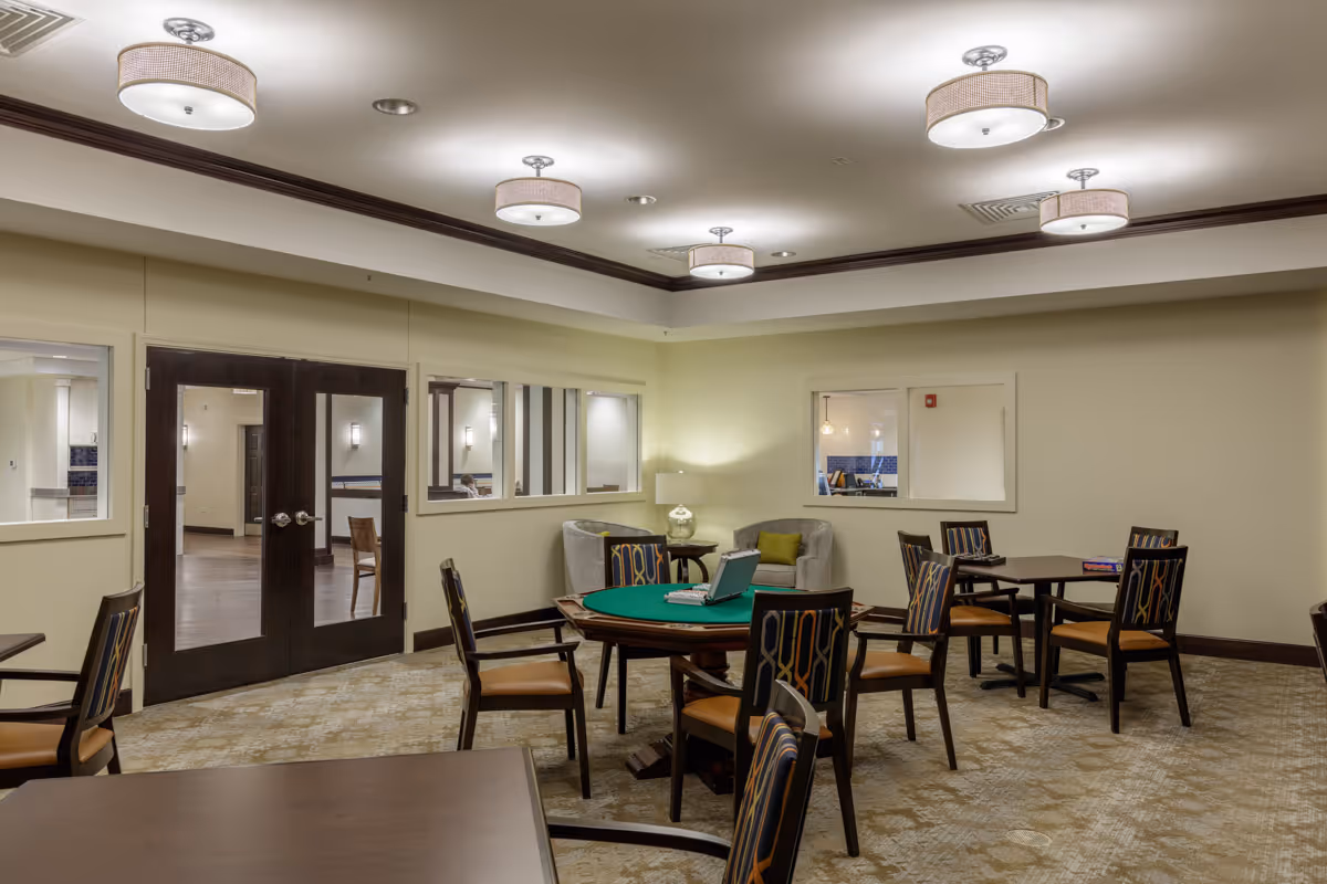 A well-lit common room with multiple tables and chairs arranged for group activities or games. One table has a green felt surface with a laptop and some items on it. The room has beige walls, carpeted floor, and modern ceiling lights. There are two armchairs with a small table and lamp in the corner, and windows looking into adjacent rooms.