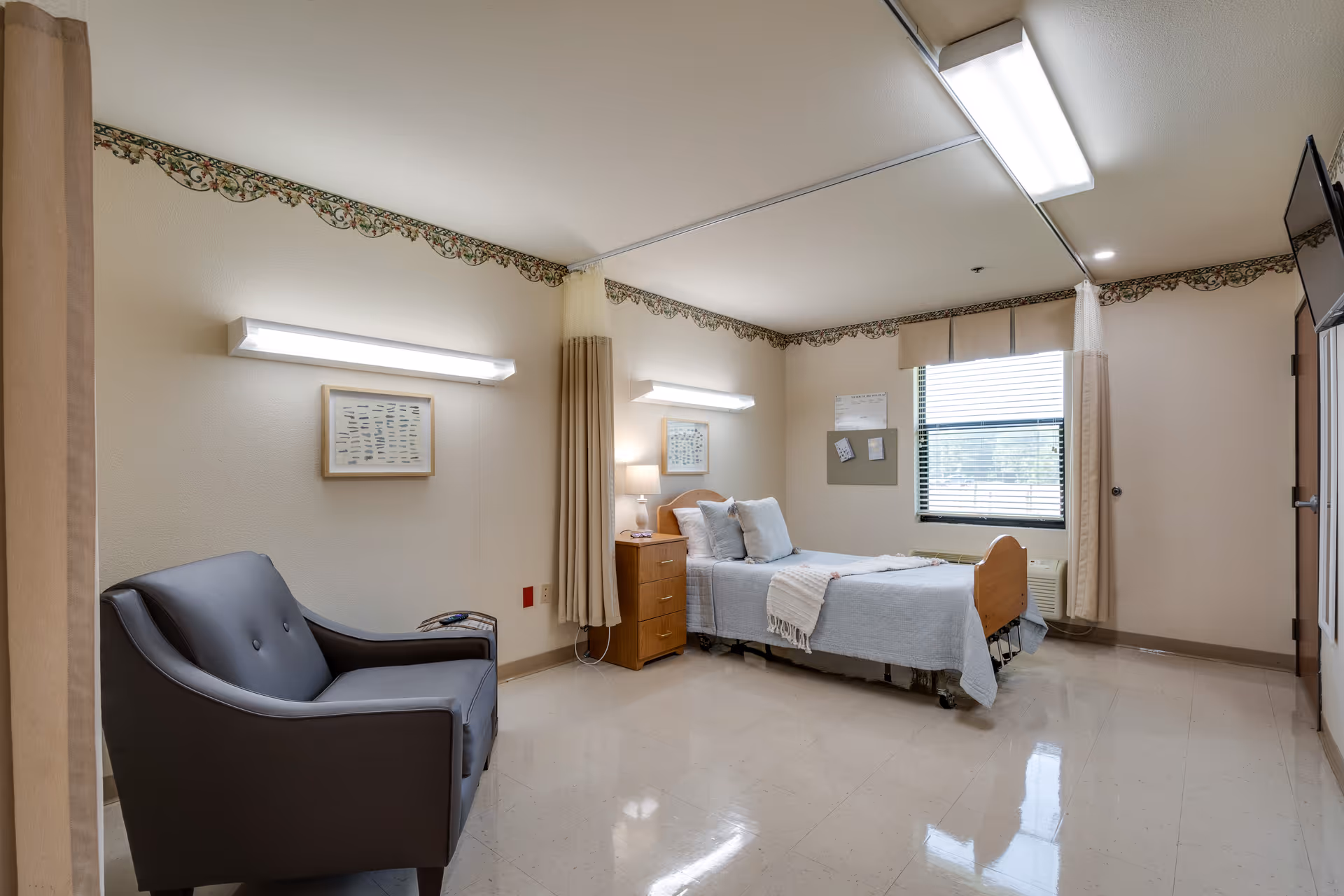 A clean and simple senior living facility bedroom with a single bed made with white linens and pillows, a wooden nightstand with a lamp, a gray armchair, a window with blinds and curtains, and fluorescent ceiling lights. The walls have a decorative border near the ceiling.