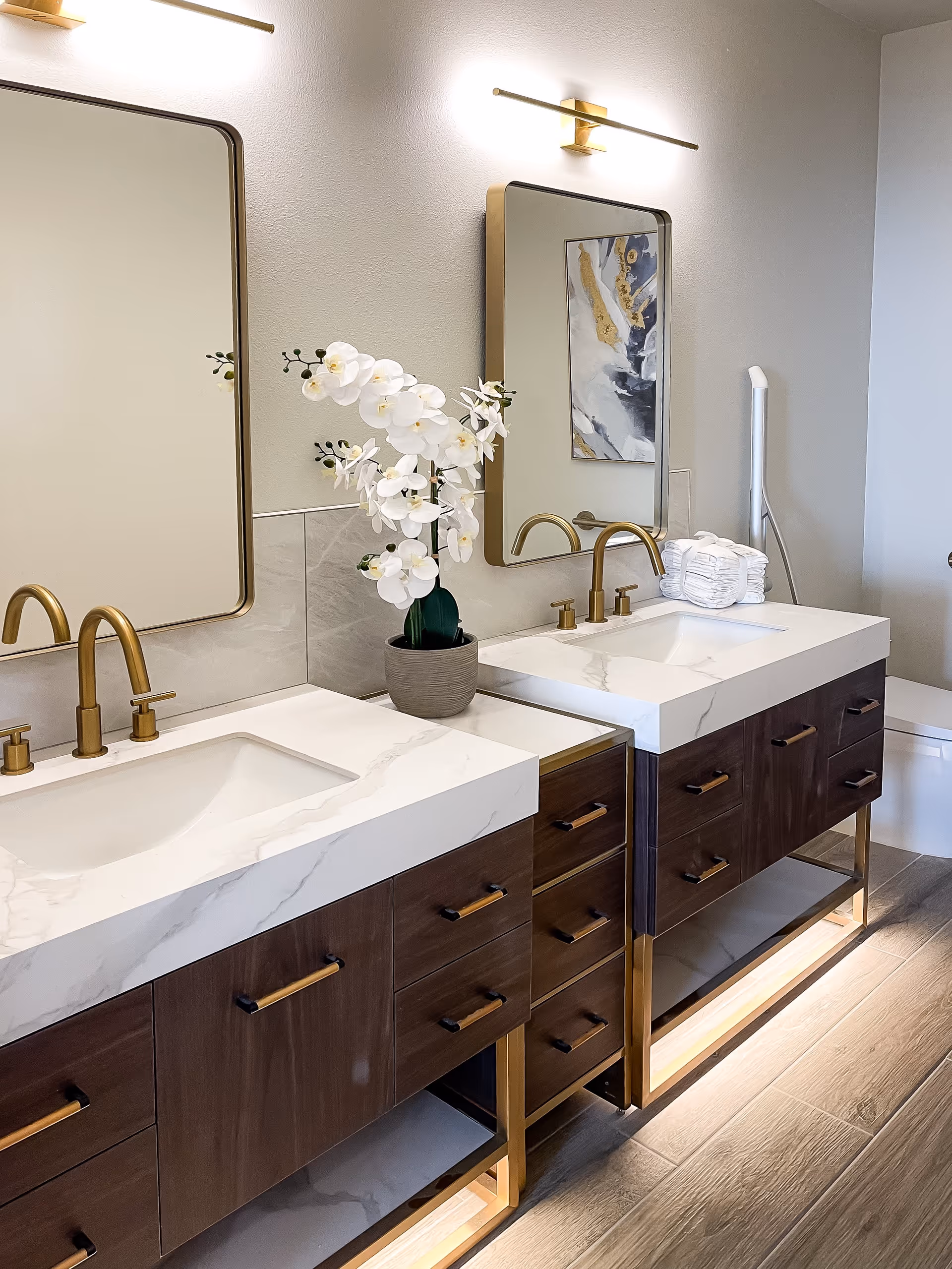 Modern bathroom with two white marble sinks set in wooden vanities with gold fixtures. Above each sink is a rectangular mirror with rounded corners and a gold light fixture. A white orchid plant in a gray pot is placed between the sinks. A stack of white towels and a toilet are visible in the background.