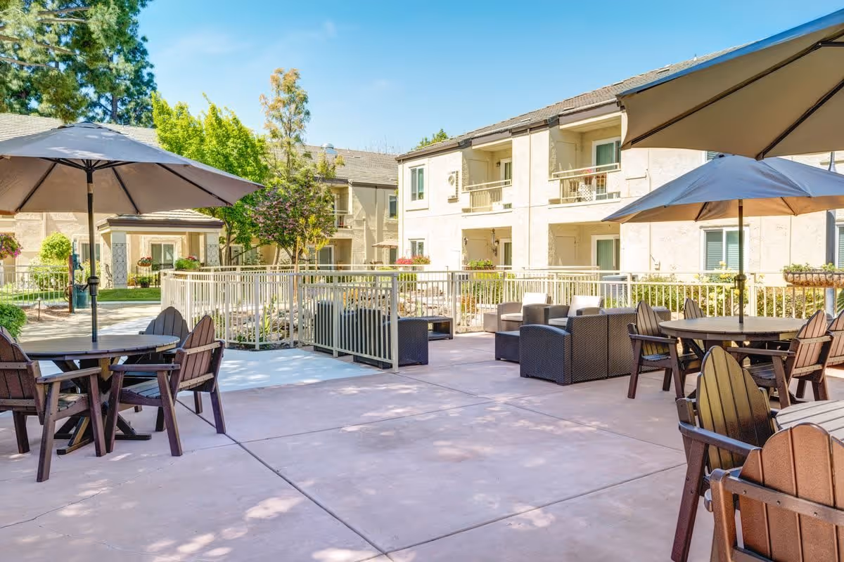 Outdoor patio area at The Oaks at Inglewood with several round tables and chairs under large umbrellas, surrounded by a two-story residential building and greenery under a clear blue sky.
