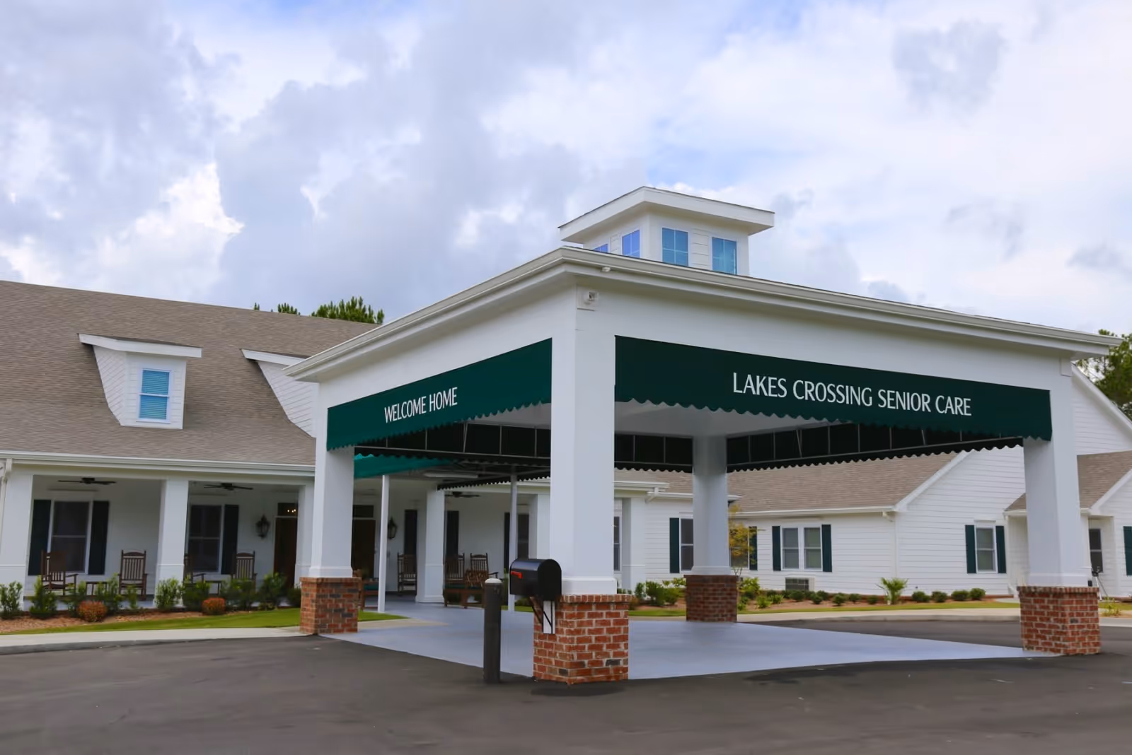 Exterior view of Lakes Crossing Senior Care facility entrance with a covered driveway supported by white pillars with brick bases. The green awning displays the text 'WELCOME HOME' and 'LAKES CROSSING SENIOR CARE'. The building is white with a grey roof and has several windows and rocking chairs on the porch.
