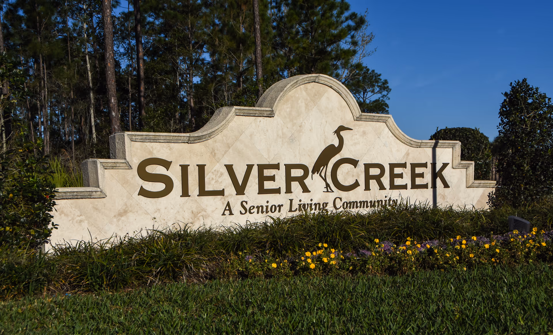 Stone sign for Silver Creek, a senior living community, surrounded by green grass, yellow and purple flowers, and trees in the background under a clear blue sky.