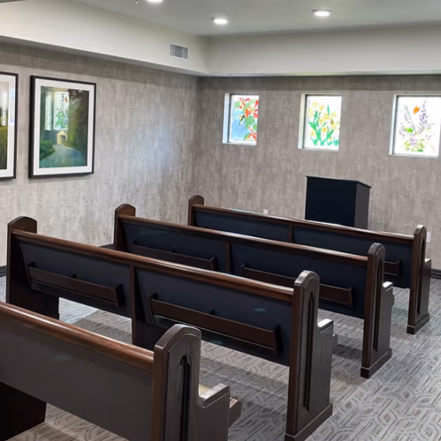 Interior view of a small chapel or meditation room with wooden pews arranged in rows facing a black podium. The walls are light-colored with three small stained glass windows depicting floral designs and two framed paintings on the left wall. The ceiling has recessed lighting.