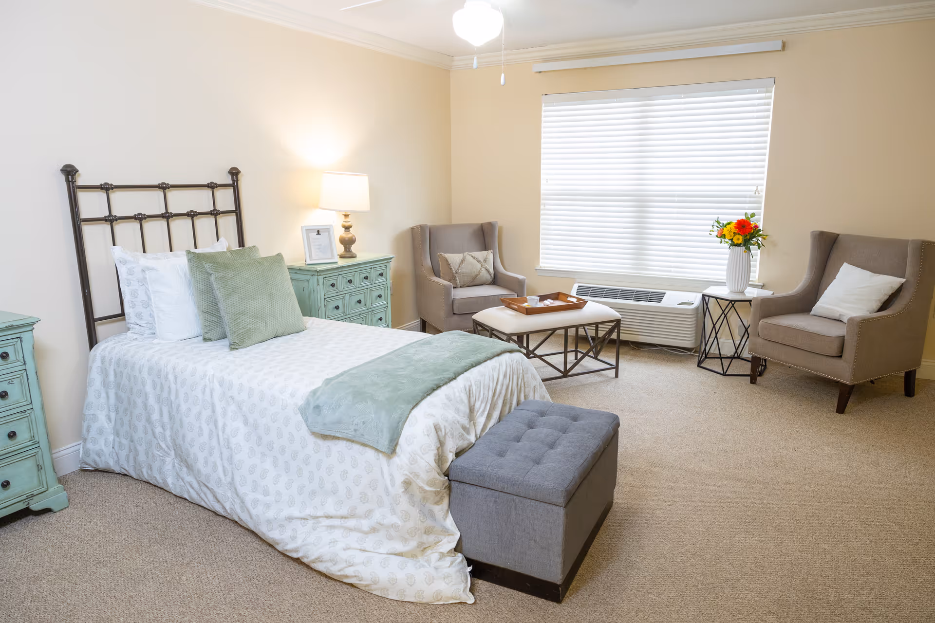 A bright and cozy bedroom with a single bed featuring white and green bedding, a green nightstand with a lamp and picture frame, two beige armchairs with cushions, a small table with a vase of flowers, a window with blinds, and a gray ottoman at the foot of the bed.