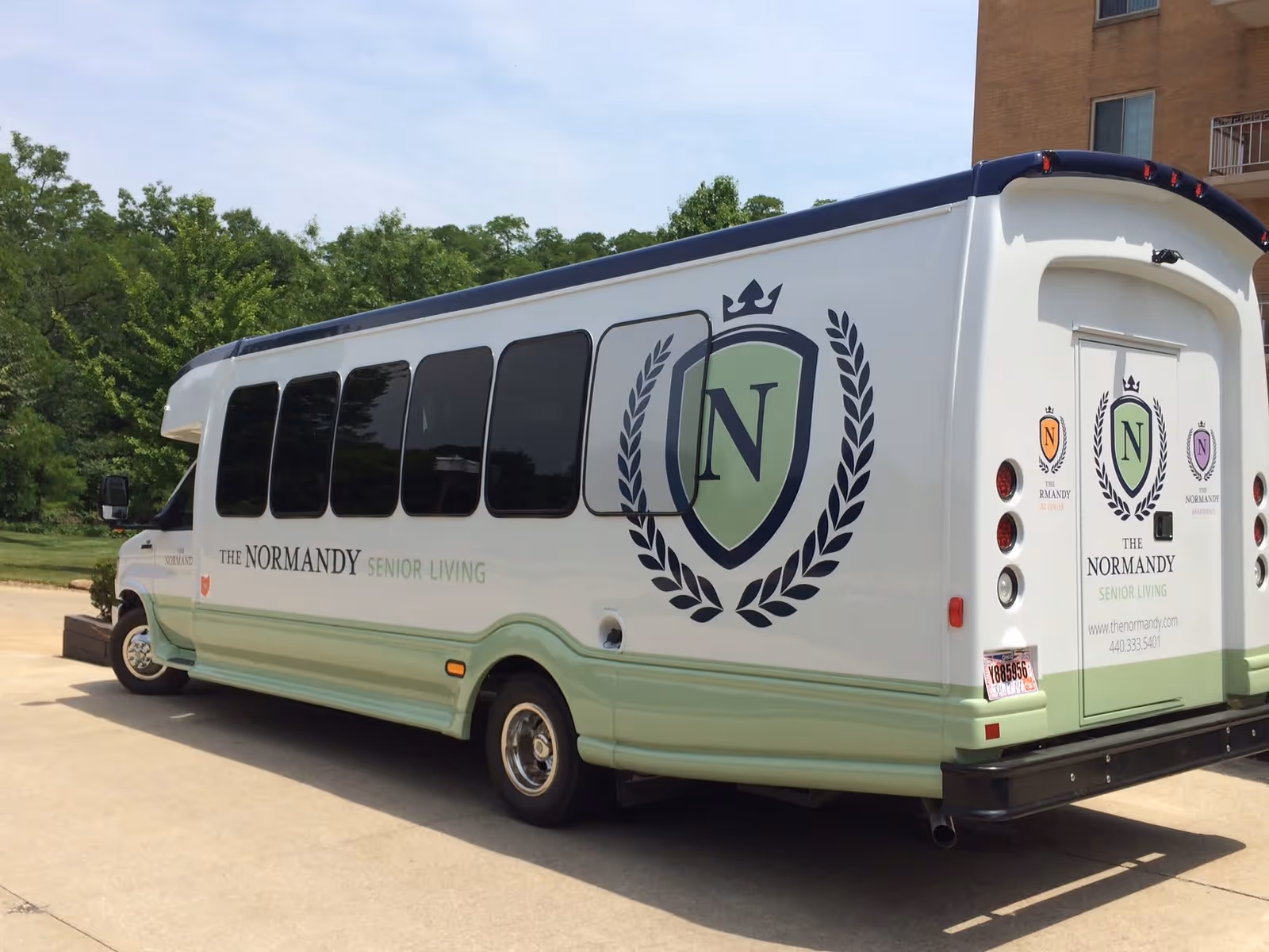 A white and light green shuttle bus parked on a driveway with large windows and logos for The Normandy Senior Living on the side and back, surrounded by trees and a building in the background.