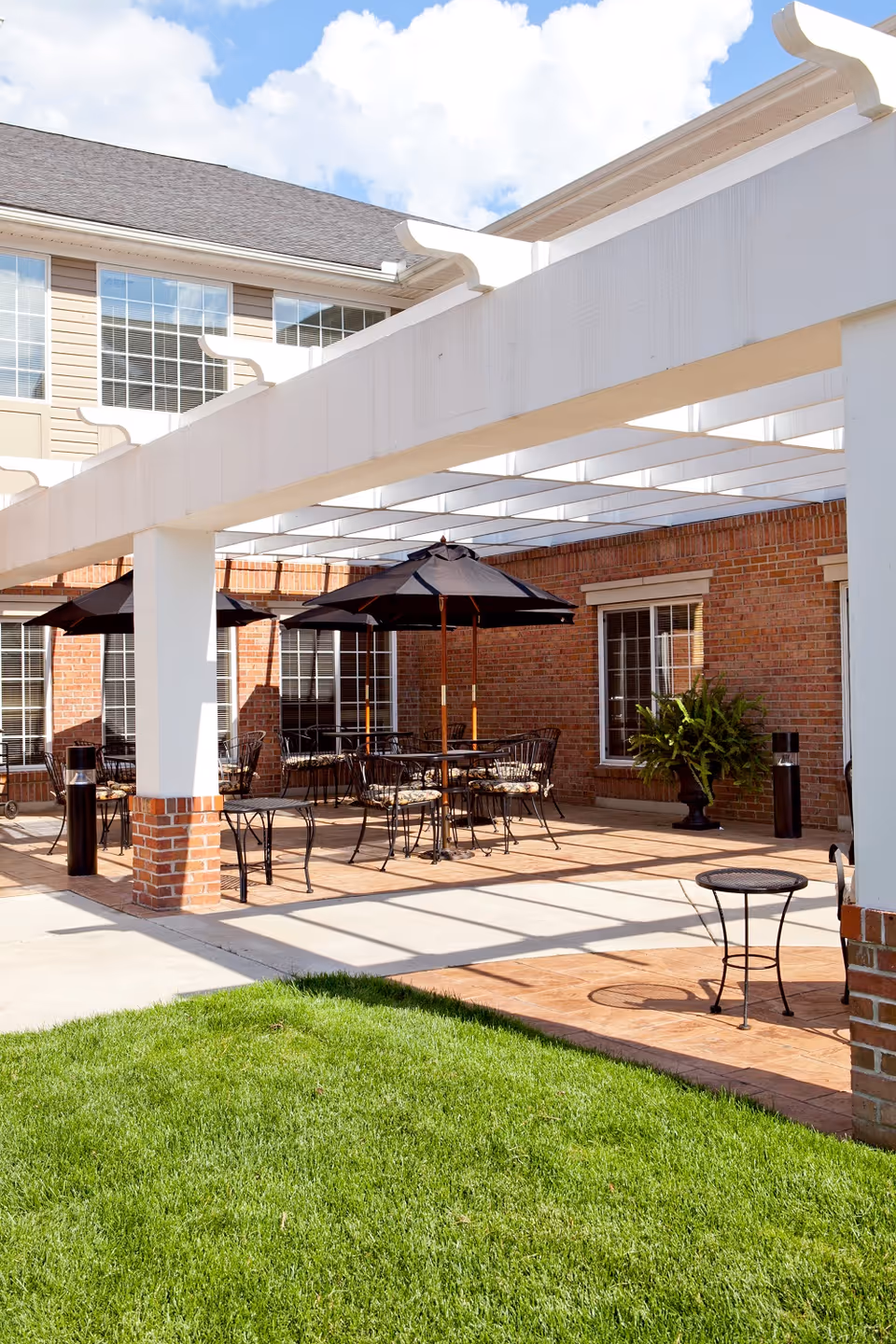 Outdoor patio with a white pergola, tables and umbrellas beside a brick building and a green lawn.