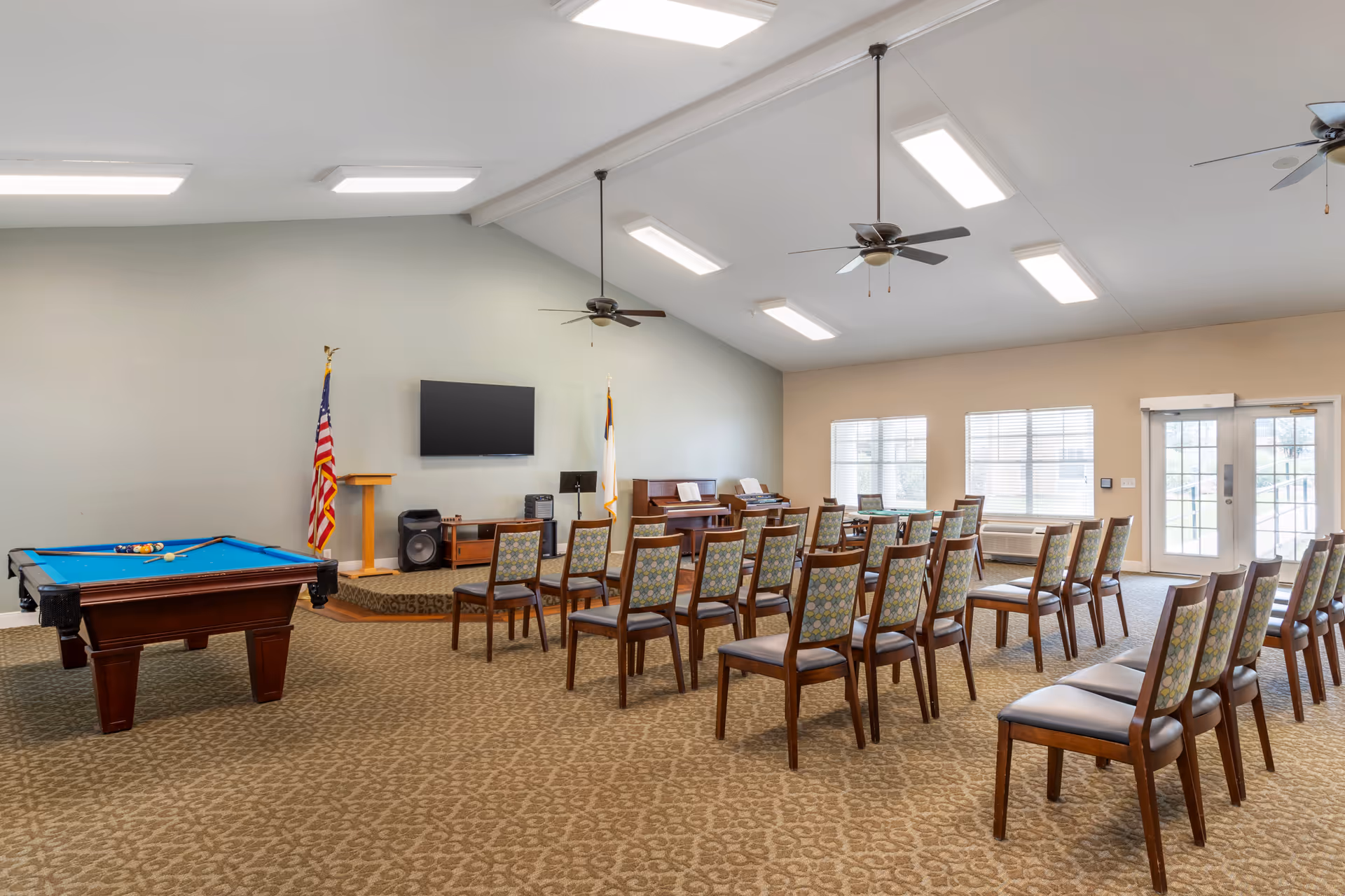 Spacious senior community activity room with rows of chairs facing a small stage, a wall-mounted TV, flags, and a pool table.