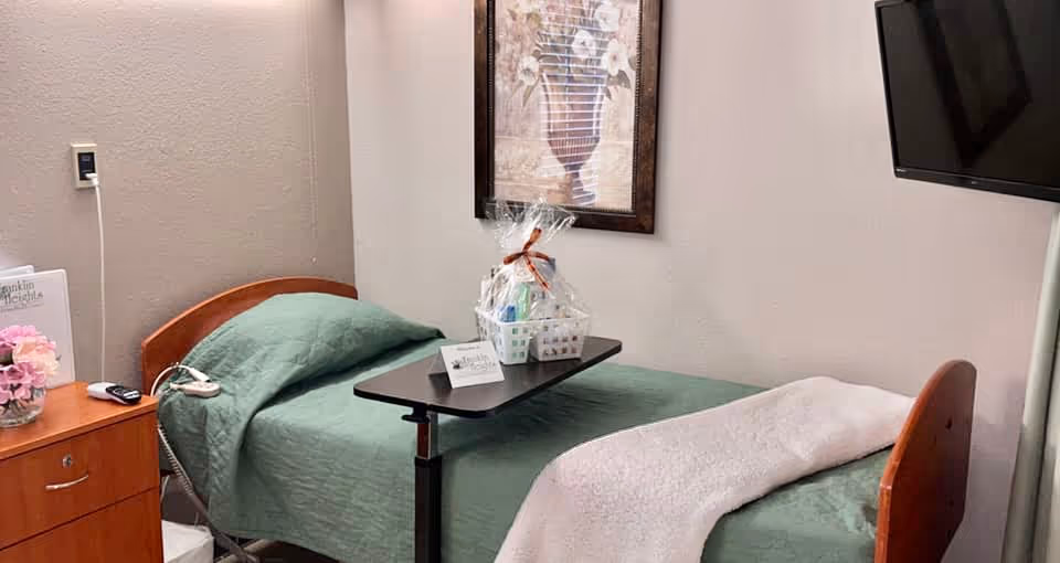 Single nursing-style bed with green bedding and an overbed table in a small patient room with a wall-mounted TV and framed artwork.