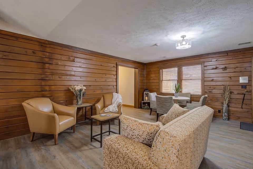 Cozy wood-paneled sitting area with a sofa, armchairs, coffee tables and a small dining nook by windows.