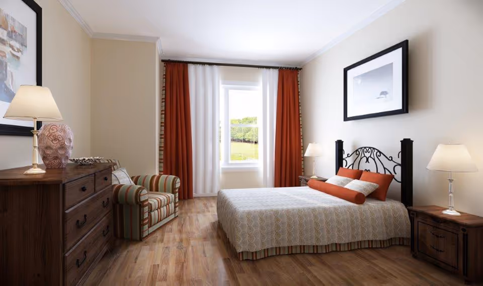 A well-lit bedroom with a large bed featuring a patterned bedspread and orange accent pillows. The room has wooden flooring, a striped armchair, a wooden dresser with a lamp and decorative vase, two bedside tables each with a lamp, and a window with white and orange curtains. A framed picture hangs above the bed.