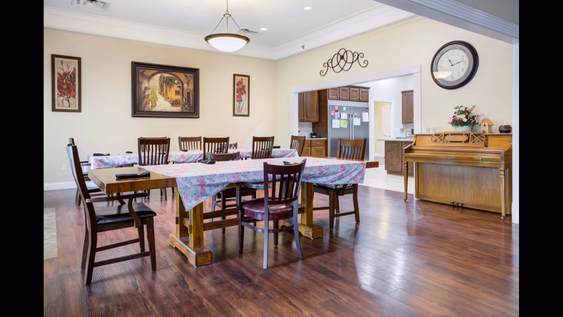 Communal dining room with wooden tables and chairs covered in floral tablecloths, a piano and a large wall clock.