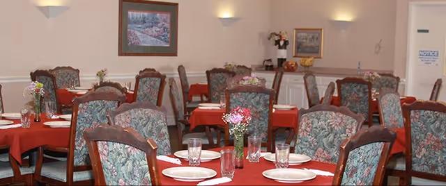 Dining room with multiple tables set with red tablecloths, floral-upholstered chairs, and place settings with vases of flowers.