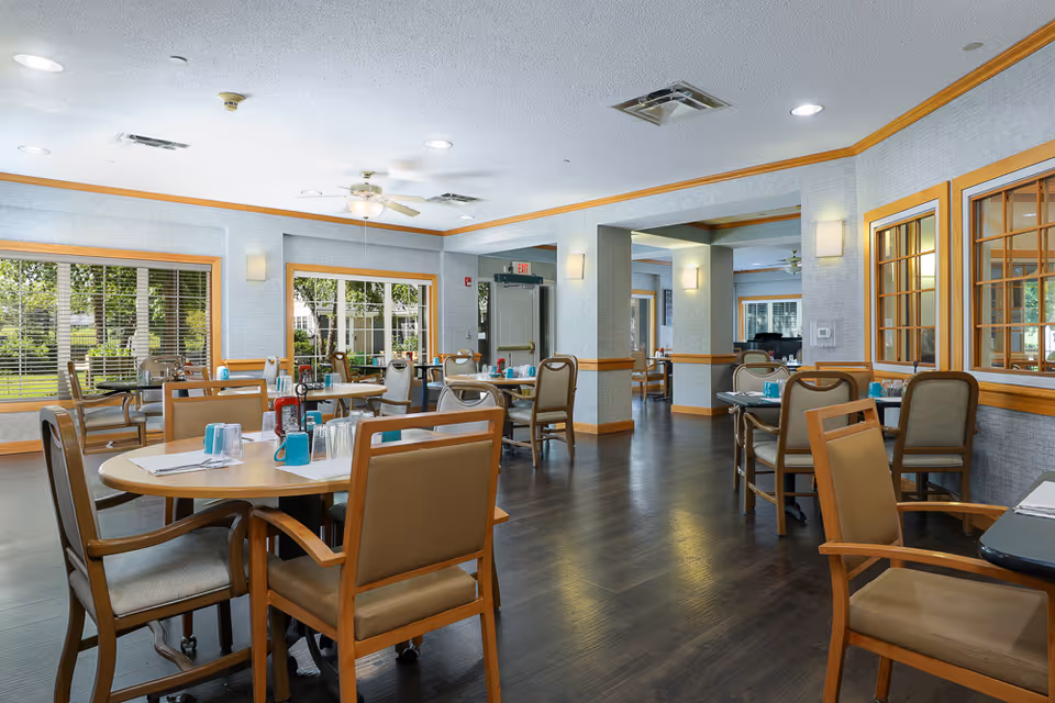 A bright and spacious dining room in an assisted living facility with multiple round and rectangular tables set with napkins, cups, and condiments. The room features large windows with views of greenery outside, wooden trim, and comfortable chairs. Ceiling fans and recessed lighting illuminate the space.