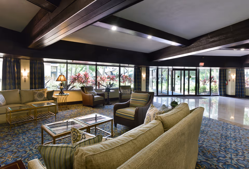 Spacious living room area with large windows letting in natural light, featuring beige sofas, patterned armchairs, glass-top coffee tables, a side table with a lamp, and a blue patterned carpet. The room has dark wooden ceiling beams and glass doors leading outside.