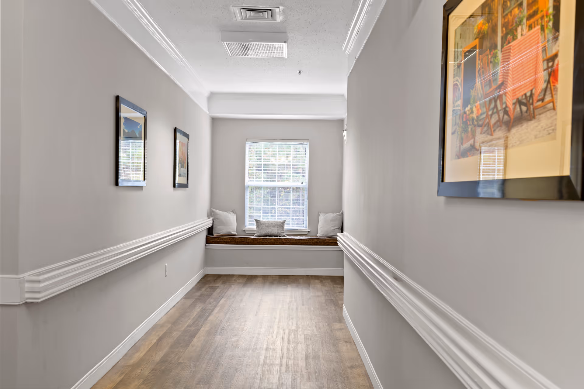 Narrow interior hallway with wood flooring leading to a window seat with cushions and framed artwork on the walls.