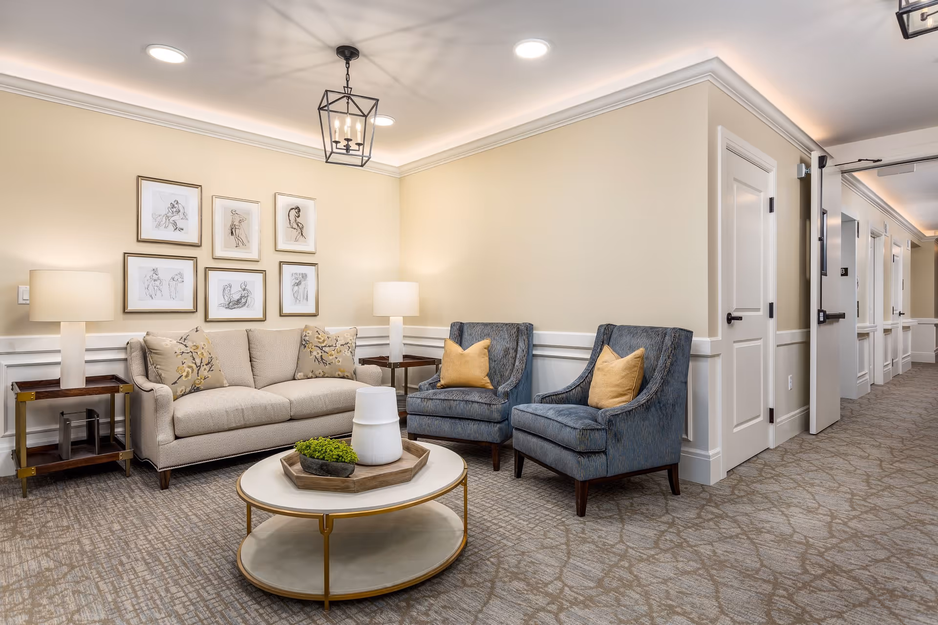 A cozy seating area in a senior living facility with a beige sofa adorned with floral pillows, two blue armchairs with yellow cushions, a round coffee table with decorative items, two side tables with lamps, framed artwork on the wall, and a hallway with multiple doors leading to rooms.