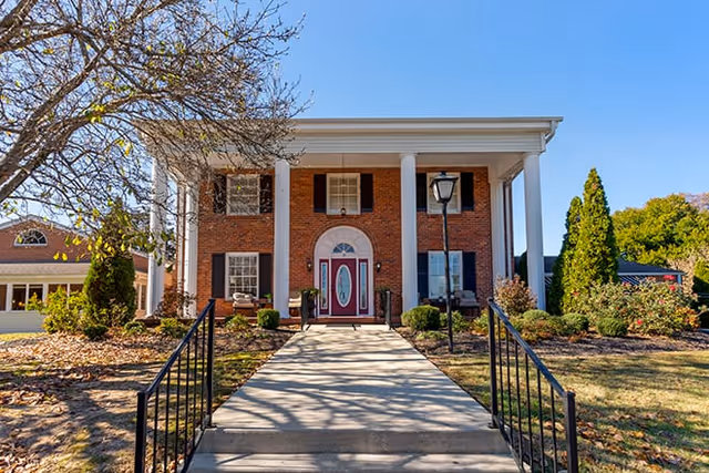 Front exterior view of a two-story brick building with white columns, a red door with an oval window, black shutters, and a concrete walkway leading up to the entrance. There are trees and shrubs around the building under a clear blue sky.