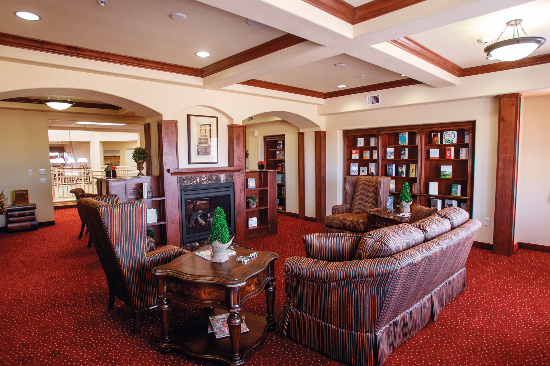 A cozy living room area in a senior living facility featuring striped upholstered sofas and armchairs arranged around a wooden coffee table with a small decorative plant. The room has a red carpet, wooden trim, built-in bookshelves filled with books, and a fireplace with a framed picture above it. The ceiling has recessed lighting and a chandelier.