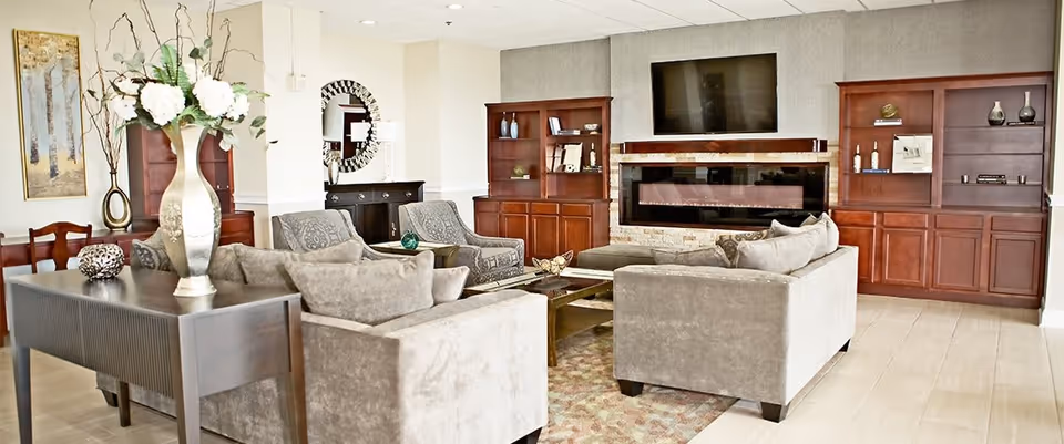 A spacious and well-lit living room area featuring two beige sofas and two armchairs arranged around a glass coffee table on a patterned rug. The room includes built-in wooden shelves and cabinets on either side of a modern electric fireplace with a large flat-screen TV mounted above it. Decorative items and vases are placed on the shelves and a console table with a large floral arrangement is positioned behind one of the sofas. A round mirror and artwork are visible on the walls.