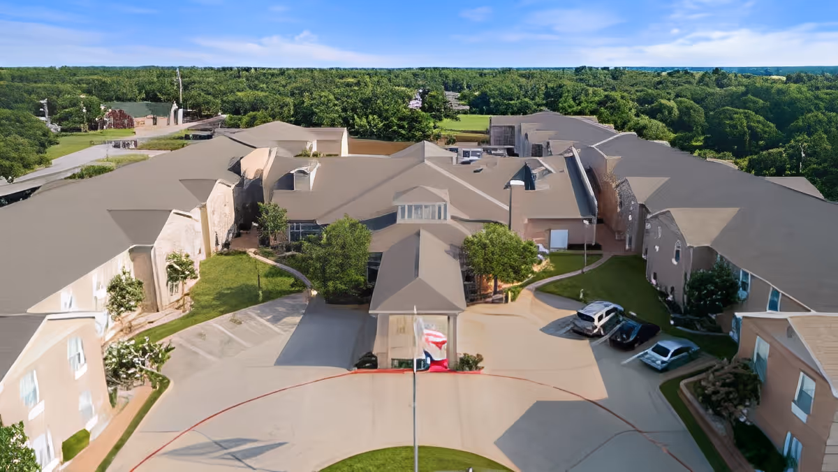 Aerial front view of a senior living complex with a circular driveway, entrance canopy, parked cars, and surrounding trees.