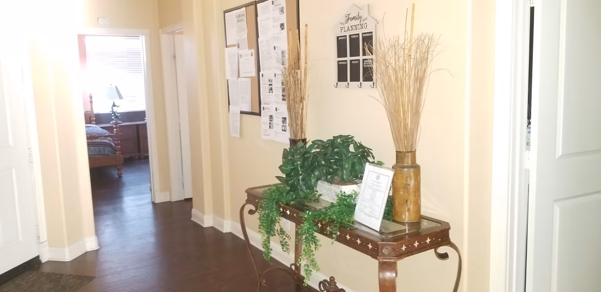Hallway interior with a decorative console table holding plants and vases, a wall bulletin board, and a doorway leading to a bedroom.