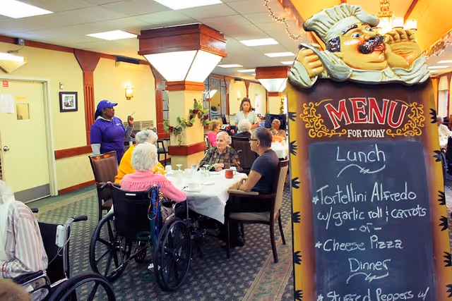 Seniors seated at a communal dining table in a dining room with staff and a large chalkboard menu.
