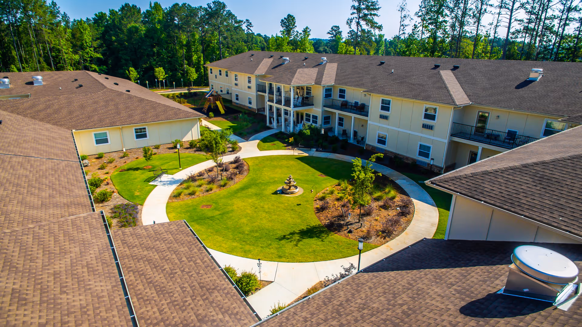 Aerial view of a senior living facility courtyard with a circular walking path, green lawn, small trees, and a central fountain. The surrounding buildings have beige walls and brown roofs, with balconies and outdoor seating areas. Trees and greenery are visible in the background.